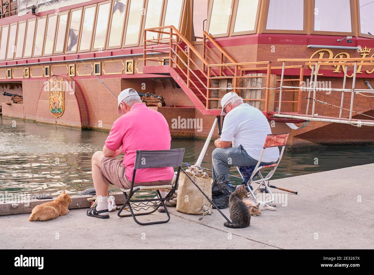 Sevastopol, Russia - January 7, 2020: Two male fishermen in casual clothes sit on folding chairs on the dock and fish. Several cute cats nearby are wa Stock Photo