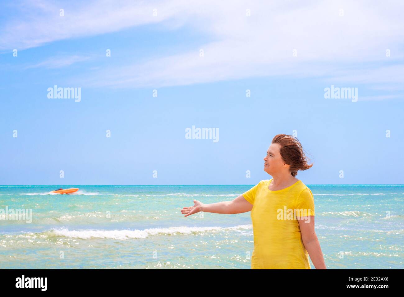 an elderly woman on the shores of the azure sea against a blue sky, stretching out her hand to the side, and enjoys a summer day, the end of quarantin Stock Photo