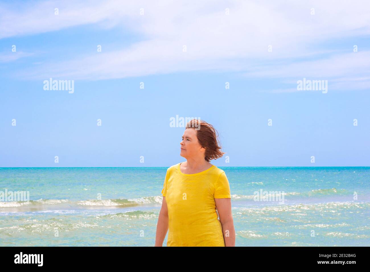 An elderly woman over 65 in a yellow T-shirt, stands on the shore of the azure sea against a blue sky, looks to the left and enjoys a summer day, the Stock Photo