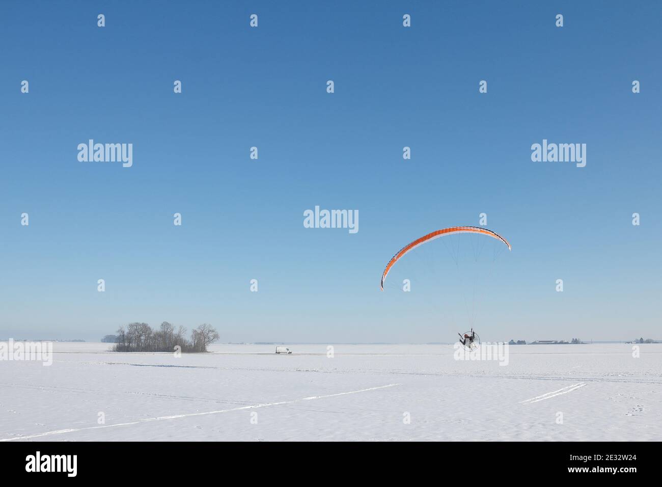 Orange motorized paraglider flying in a snowy field in winter with horizon and blue sky Stock Photo