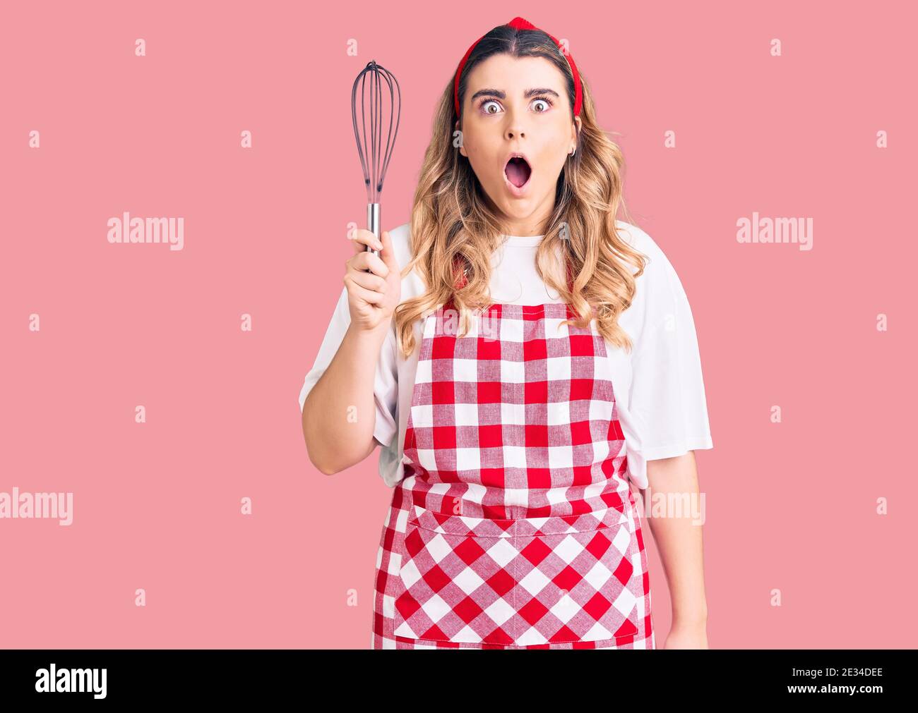 Young caucasian woman wearing apron holding whisk scared and amazed