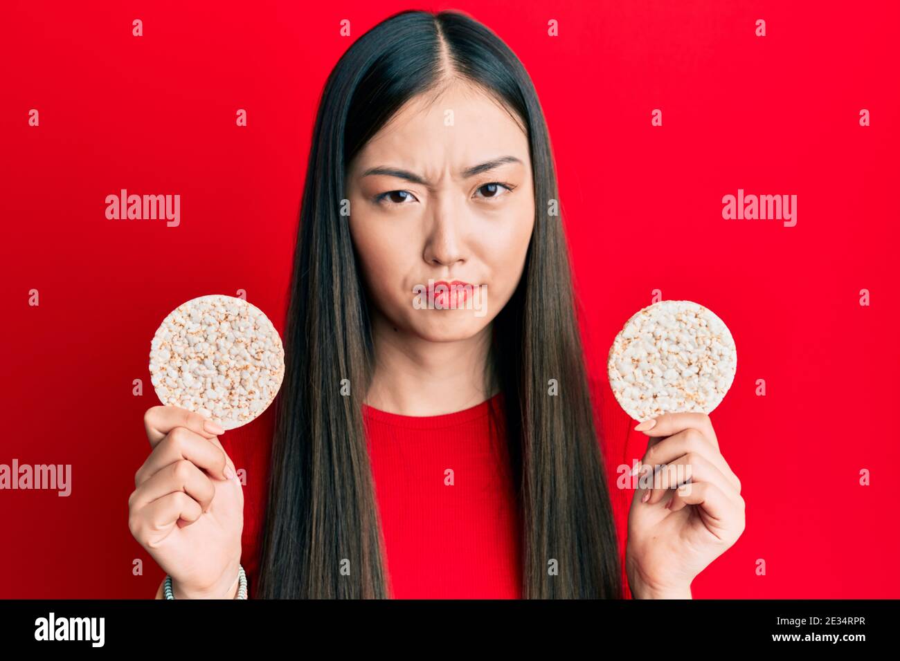 Young chinese woman eating healthy rice crackers skeptic and nervous