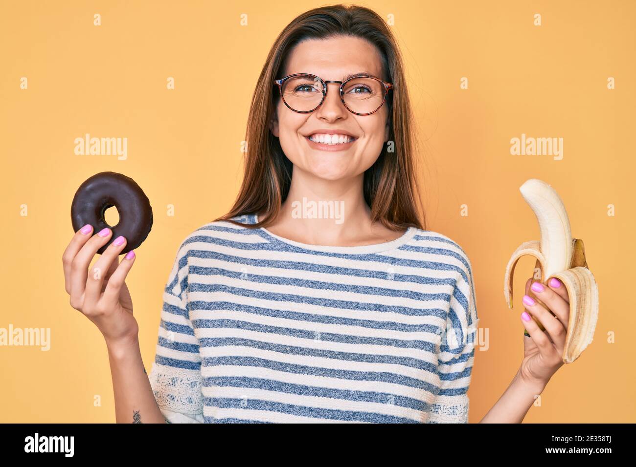 Beautiful caucasian woman holding banana and chocolate doughnut smiling with a happy and cool smile on face. showing teeth. Stock Photo