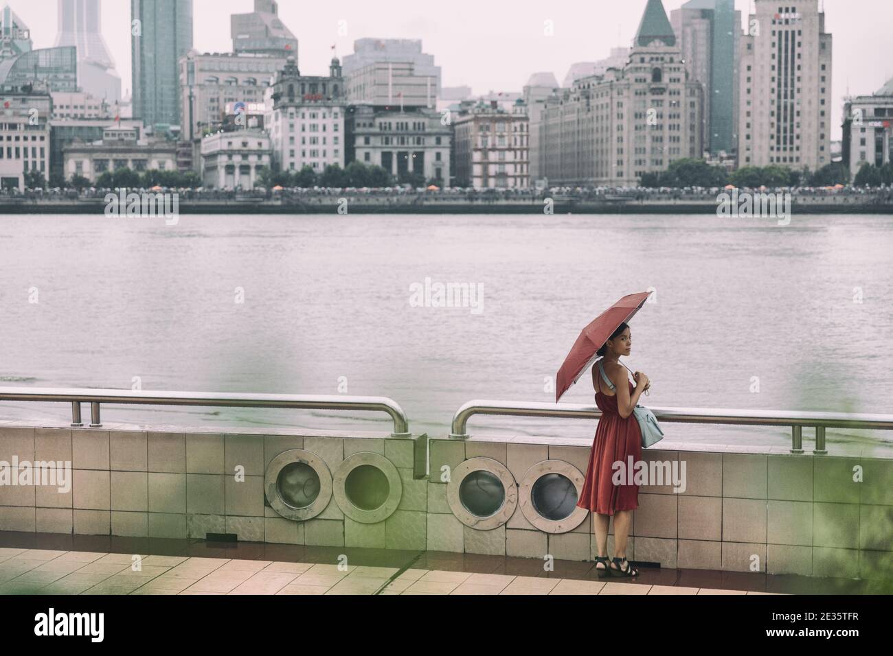 Shanghai city bund river Asian woman walking under rain with umbrella on China Asia vacation. Elegant chinese tourist lady in red dress Stock Photo