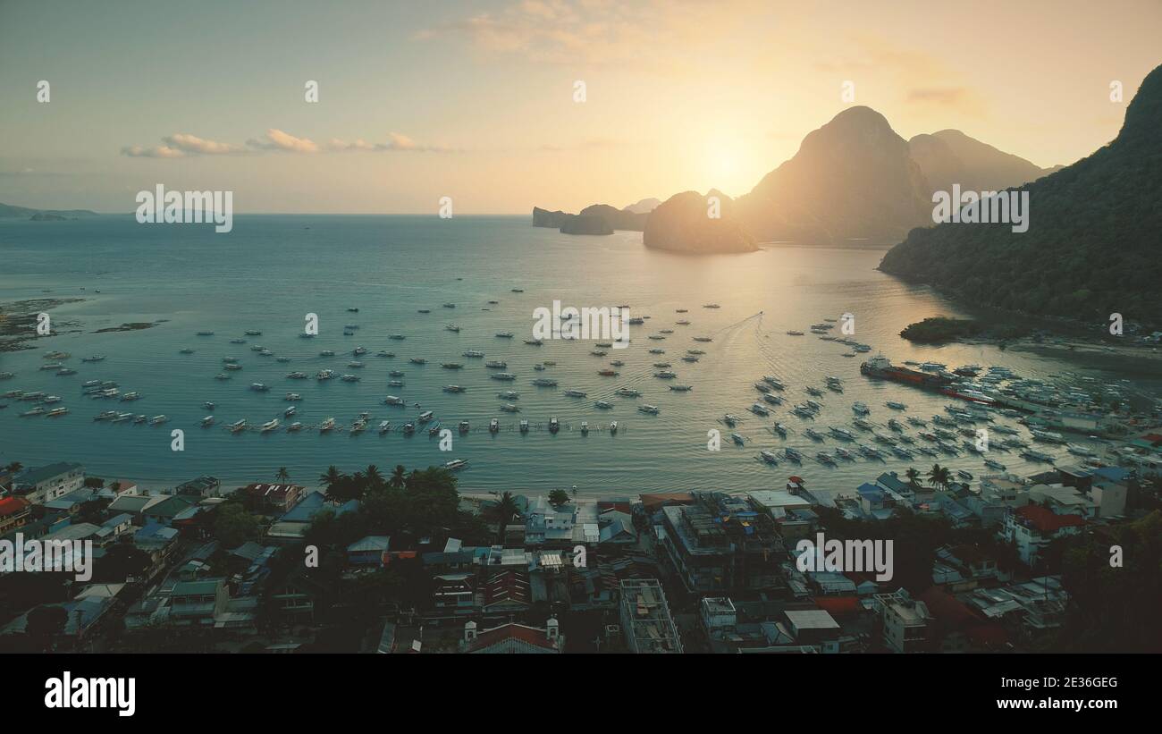 Aerial sunset at ocean harbor with boats of tropic port town cityscape. Buildings, lodges, homes with resort at sand coast. Mount Philippines island silhouette at sun light. Cinematic drone shot Stock Photo