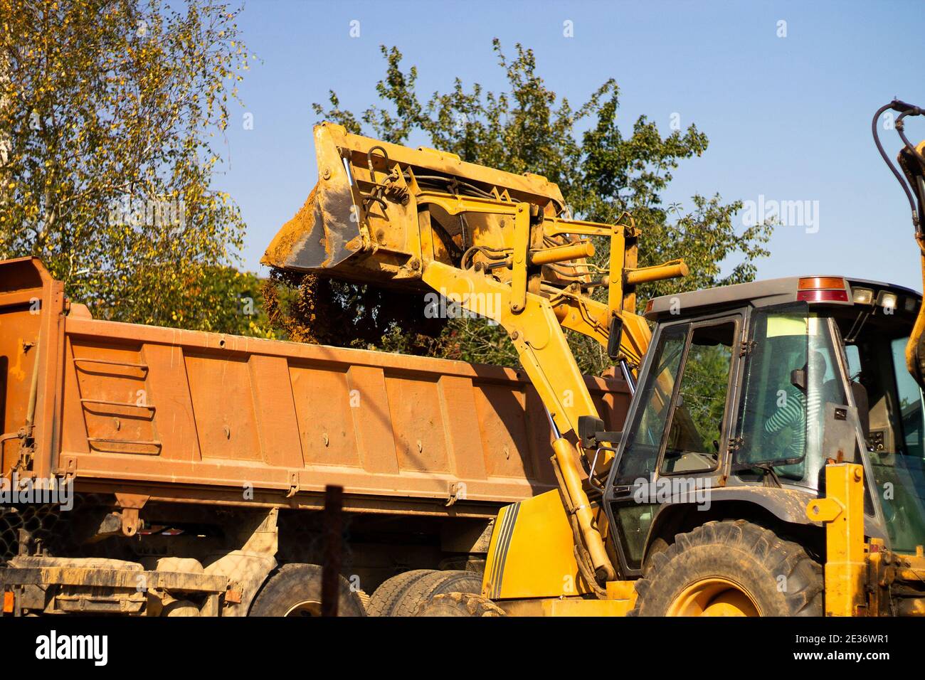 A wide metal excavator bucket digs sand or clay, loading it into a dump