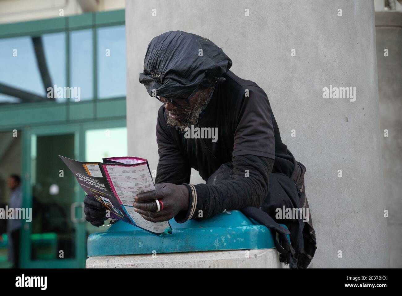 A black man with glasses, in dirty black clothes, reads a ComicCon booklet in front of the LA Convention Center leaning on trash bin Stock Photo