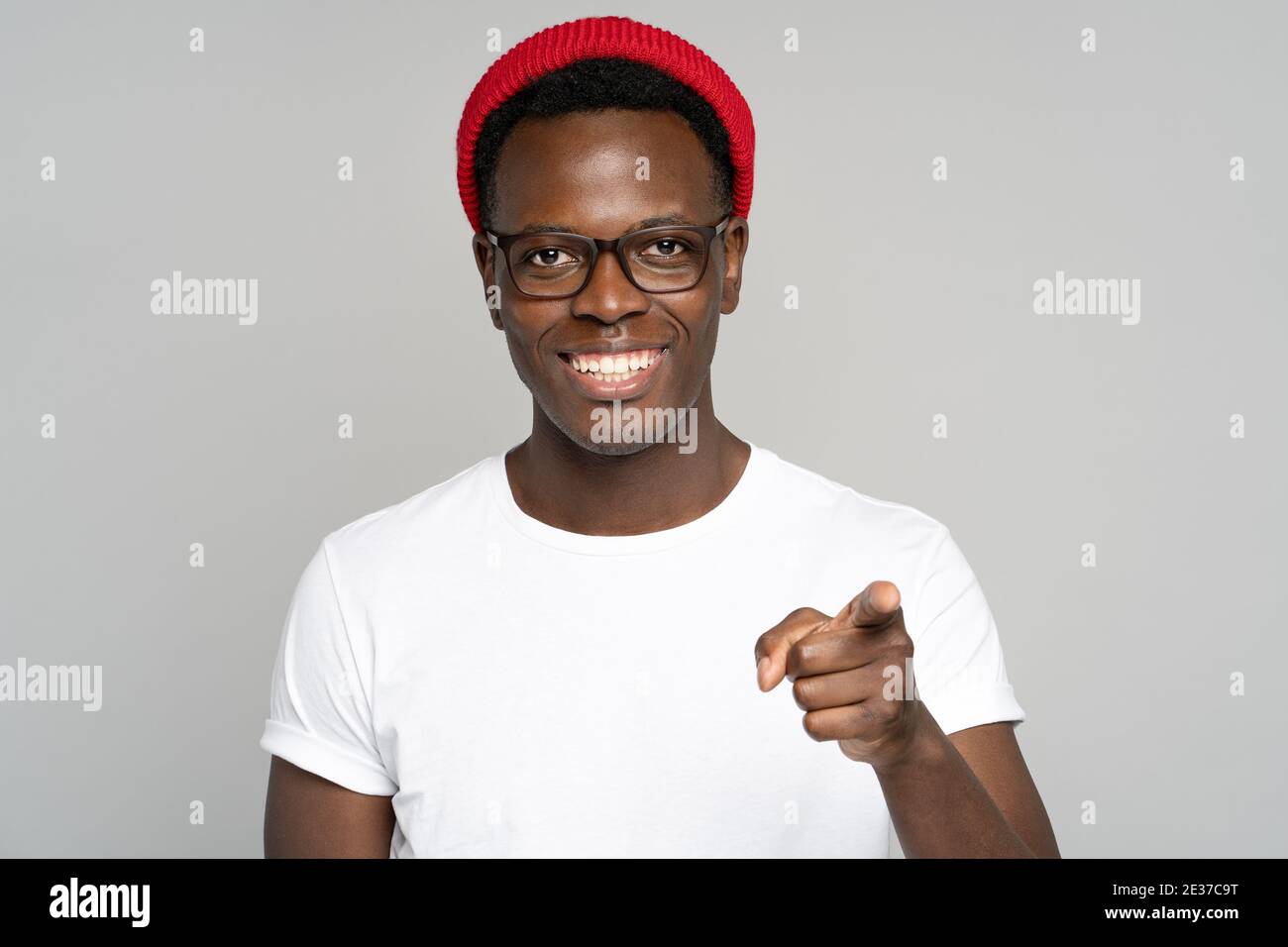 Cheerful positive Afro hipster man smiling broadly, pointing a finger at you, studio grey background Stock Photo