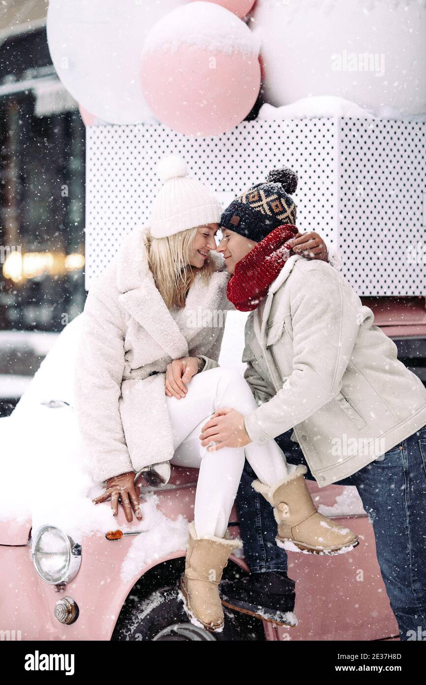 Romantic couple kissing, hugging, having fun in winter weather sitting on pink car during snowfall Stock Photo