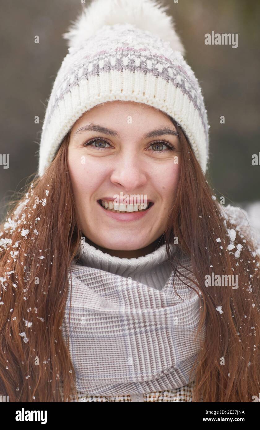 Vertical close up portrait of smiling young woman in winter wearing knit hat while looking at camera covered in snow Stock Photo