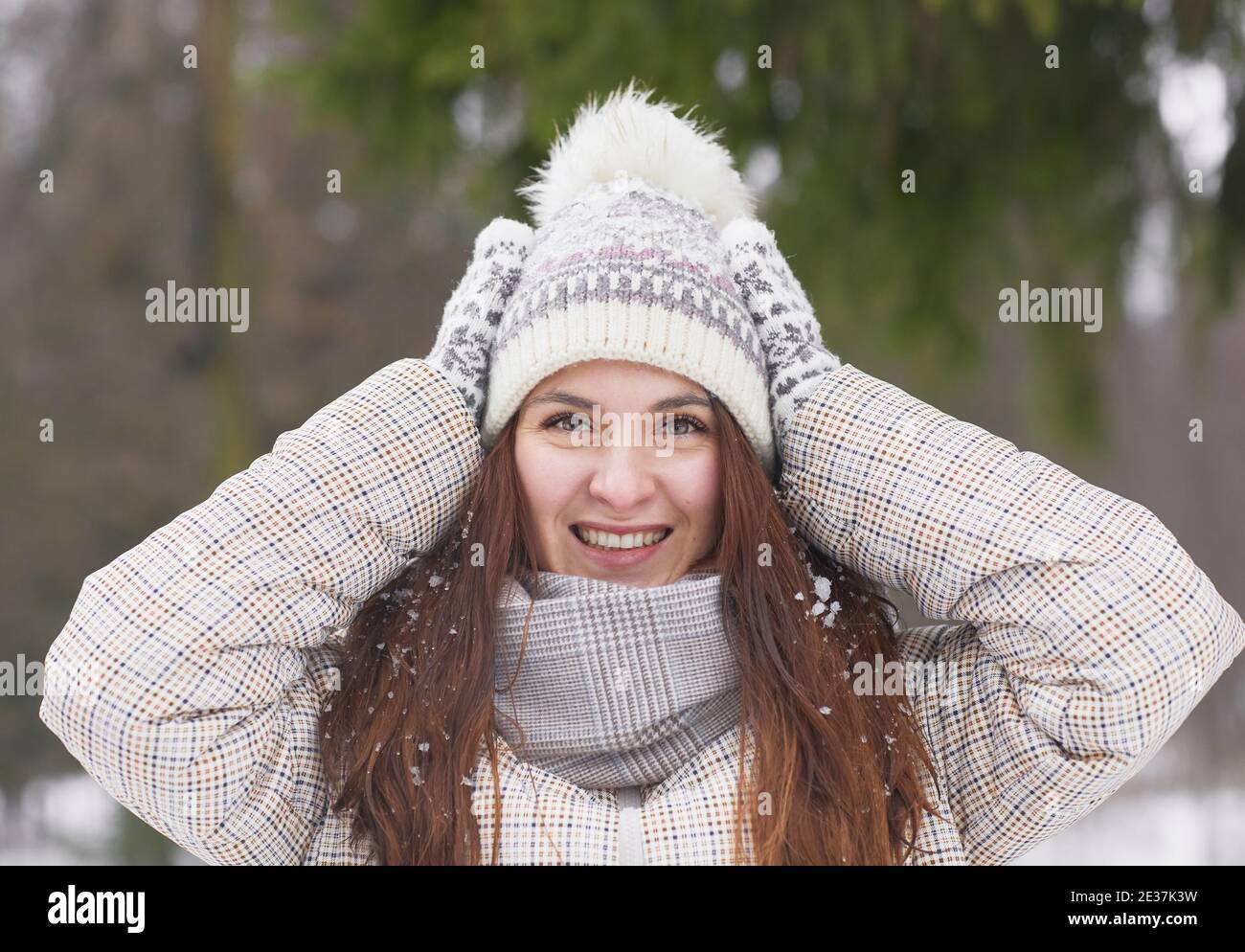 Front view portrait of happy young woman in winter outdoors wearing knit hat and mittens while smiling at camera covered in snow Stock Photo