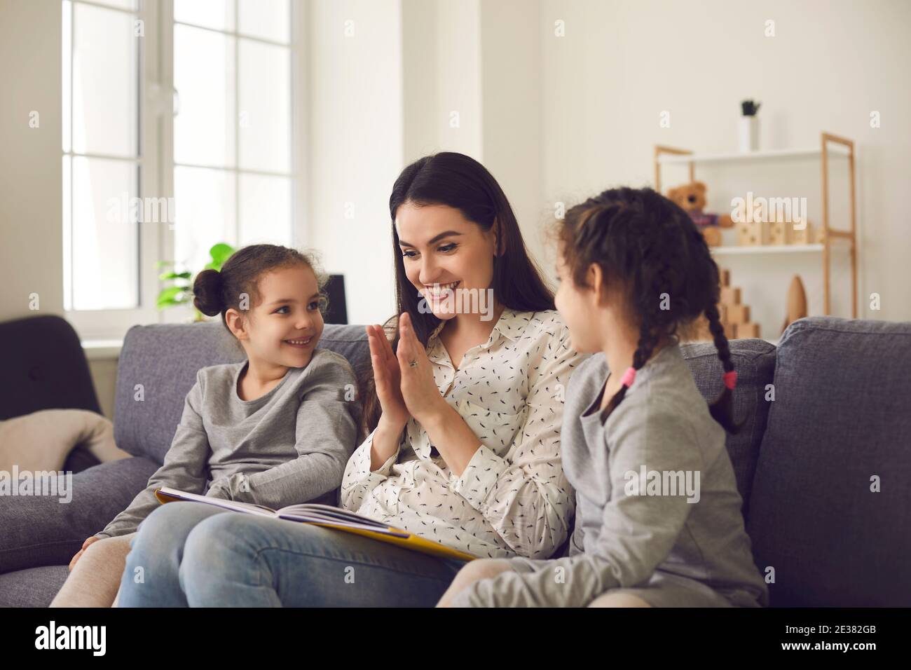 Caring smiling mom reading a book with her two little daughters sitting on the couch at home. Stock Photo