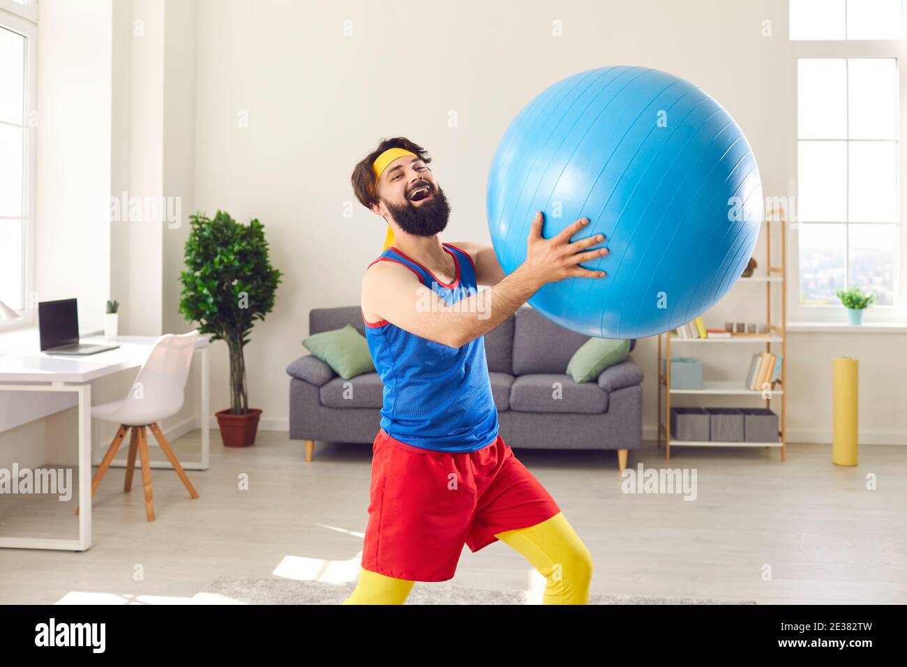 Man in colorful sportswear having fun and lifting a fitness ball during sports training at home. Stock Photo