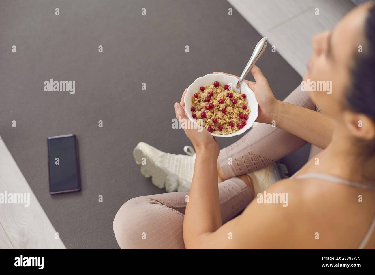 Bowl of oatmeal with cranberries in the hands of an unknown sports woman sitting on a gray yoga mat. Stock Photo