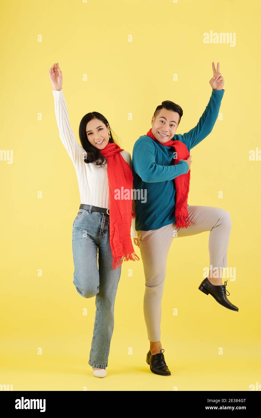 Excited happy young Asian couple in wool winter scarf dancing together against yellow background Stock Photo