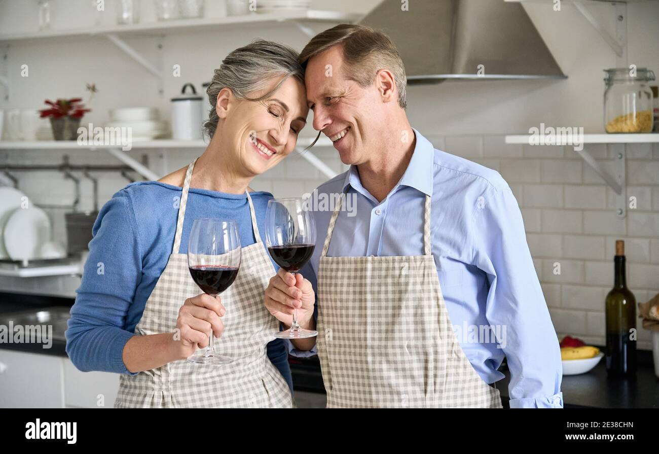 Happy old couple bonding, drinking wine standing in kitchen cooking at home. Stock Photo