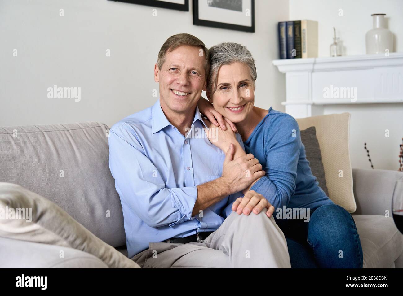 Happy older couple hugging, looking at camera, sitting on couch in apartment. Stock Photo