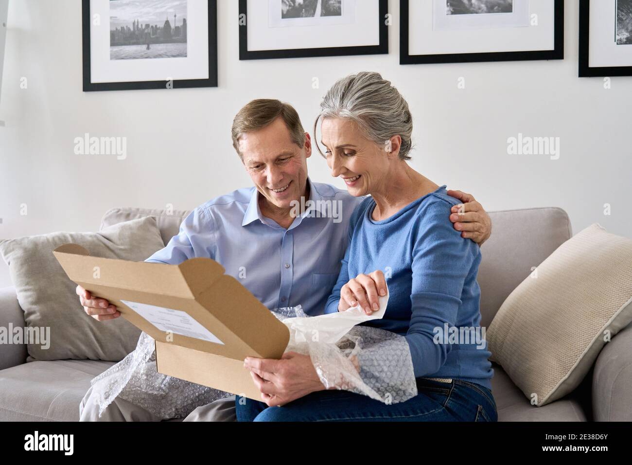 Happy older mature couple customers unpacking parcel sitting at home on couch. Stock Photo
