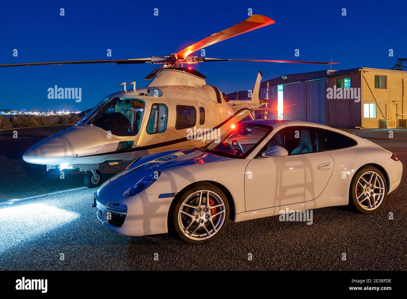 An Agusta 109 Power executive helicopter at twilight alongside a Porsche 911 car. Stock Photo