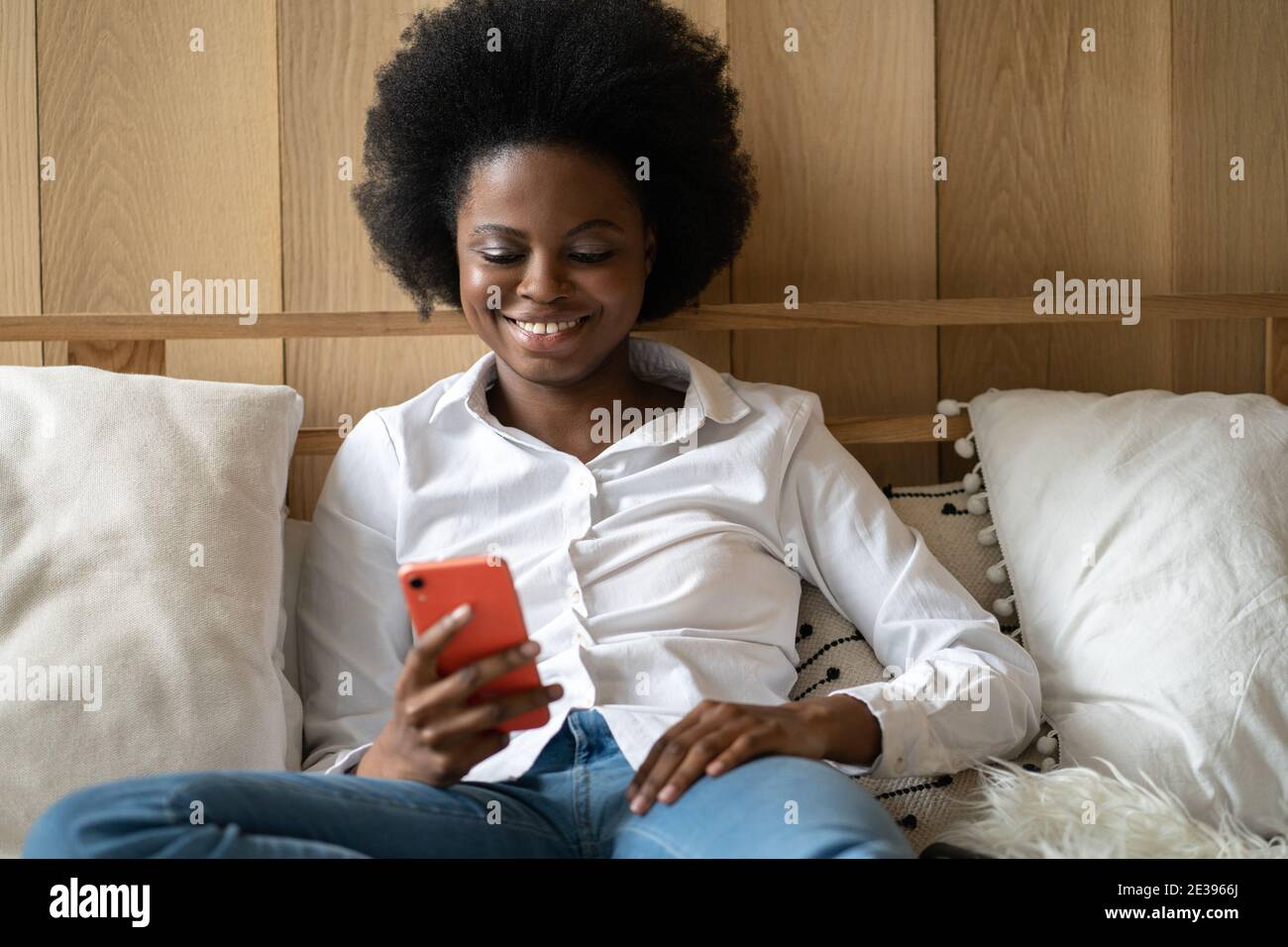 Cheerful Afro millennial woman lying on bed at home, resting, using mobile phone, typing message Stock Photo