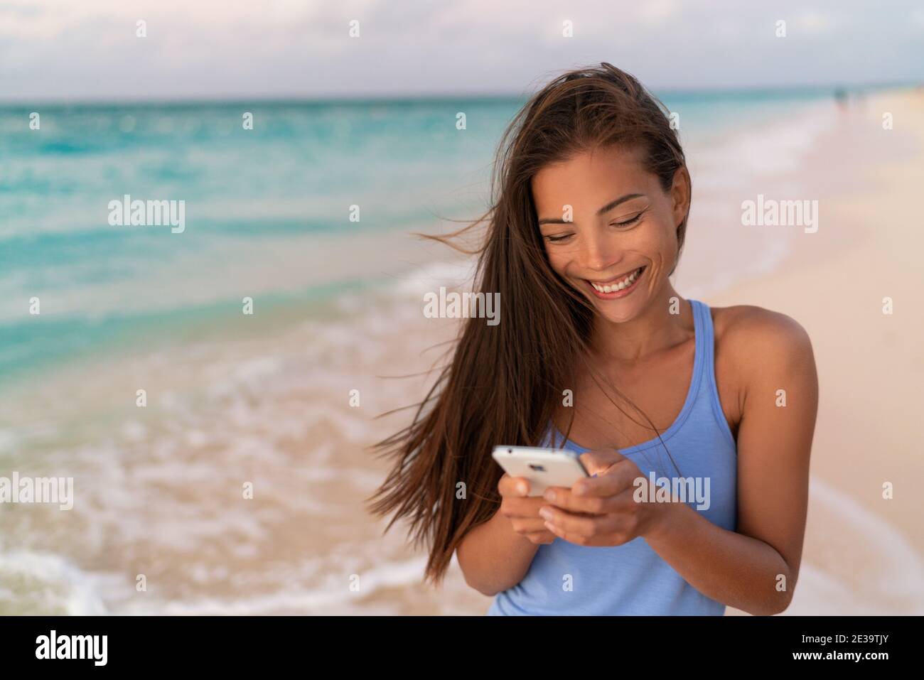 Asian woman using smart phone texting online on dating app holding cellphone mobile phone outside walking on beach at sunset. Happy young eurasian Stock Photo