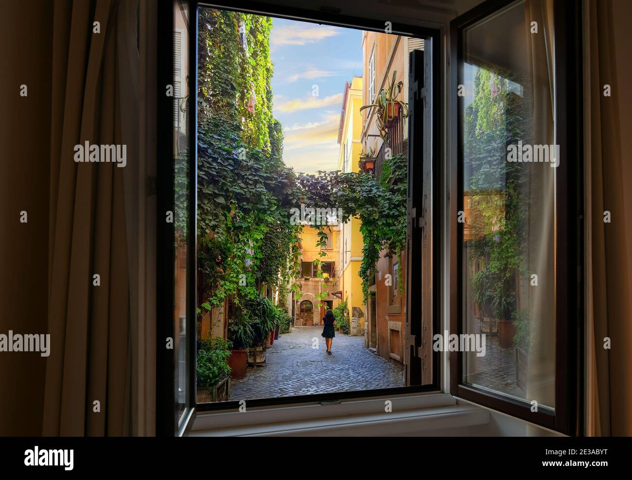 View through an open window as a young woman walks through an ivy covered colorful alley in the Trastevere district of Rome, Italy Stock Photo