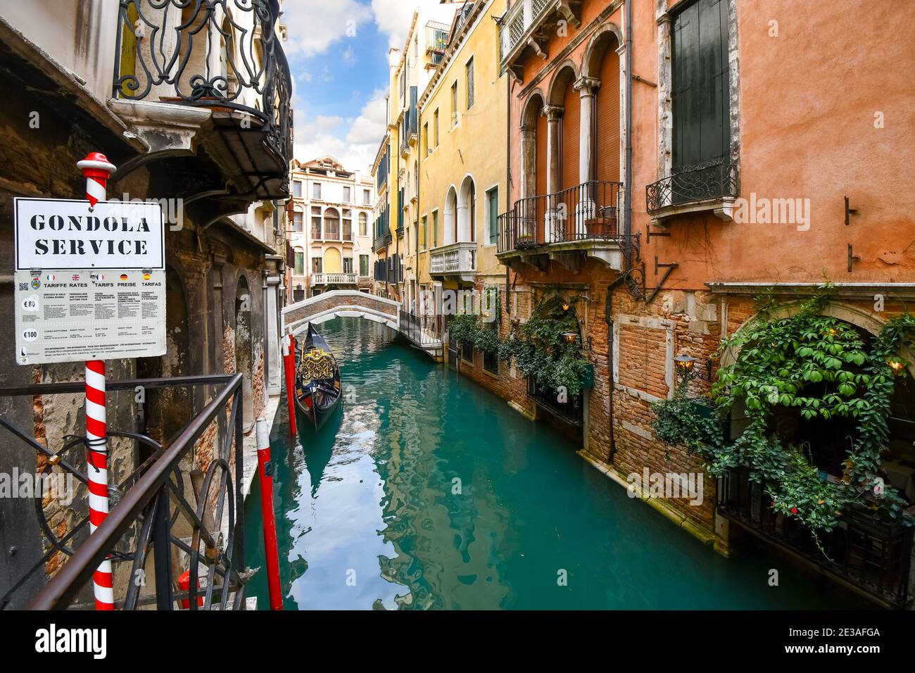 An empty gondola waits for tourists at a station on a colorful, narrow alley with a bridge and price sheet in Venice, Italy. Stock Photo