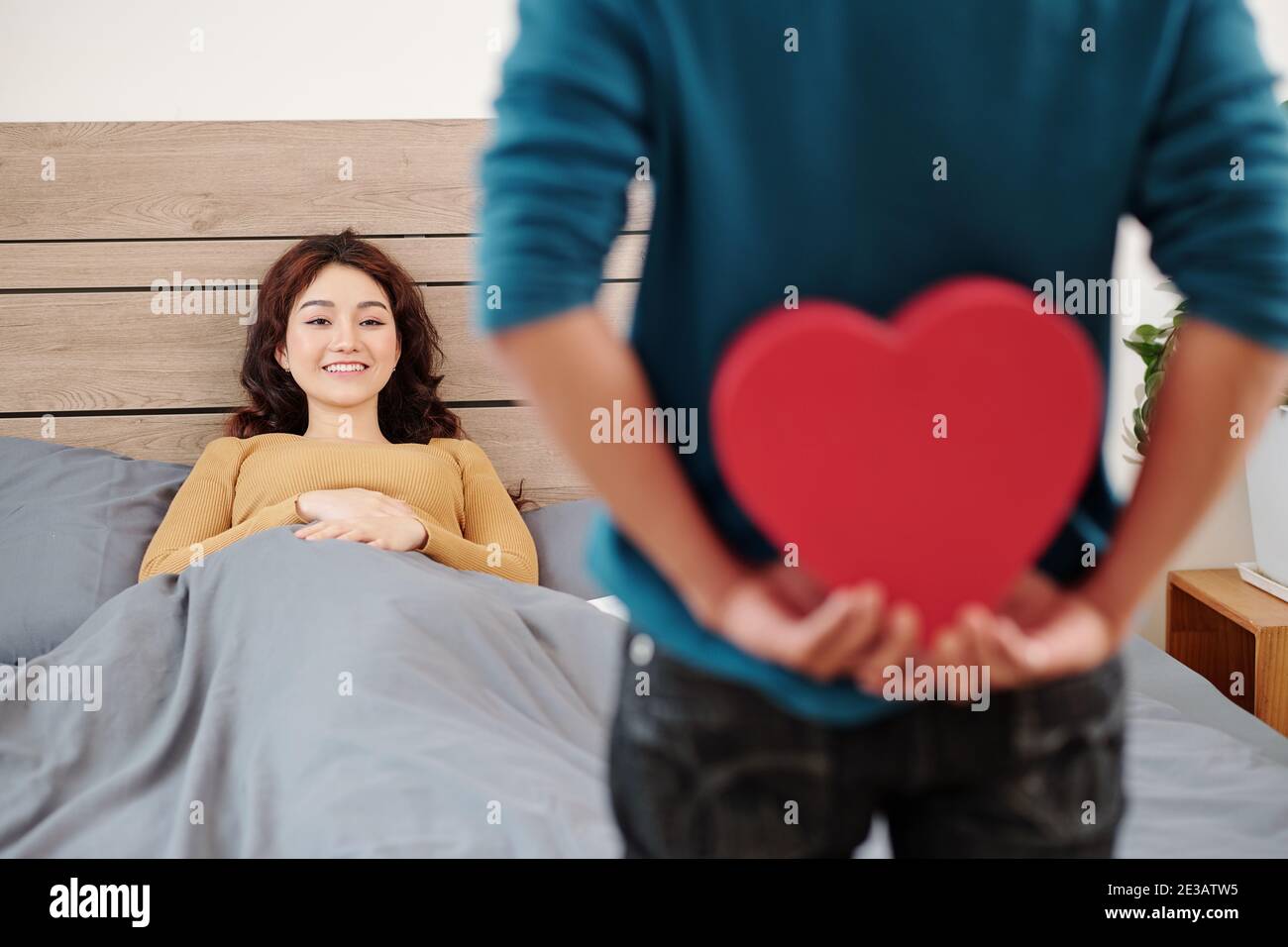 Young man hiding romantic Valentines day present for his girlfriend snuggling in bed after waking up Stock Photo