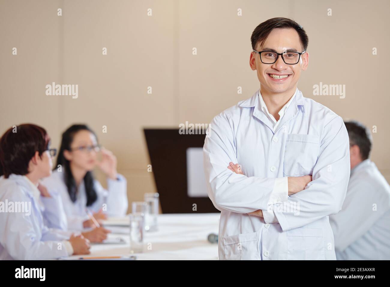 Portrait of happy smiling young scientist in labcoat standing at meeting table with arms folderd and looking at camera Stock Photo