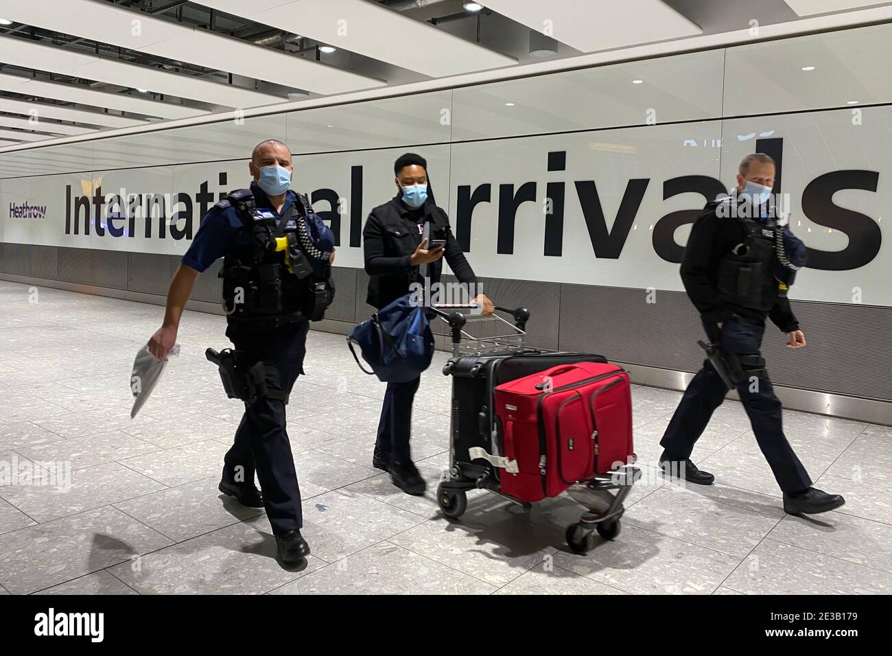 A passenger walks with police officers while pushing a trolley through the Arrival Hall of Terminal 5 at London's Heathrow Airport after arriving into the UK following the suspension of the travel corridors. Passengers arriving from anywhere outside the UK, Ireland, the Channel Islands or the Isle of Man must have proof of a negative coronavirus test and self-isolate for 10 days. Picture date: Monday January 18, 2021. Stock Photo