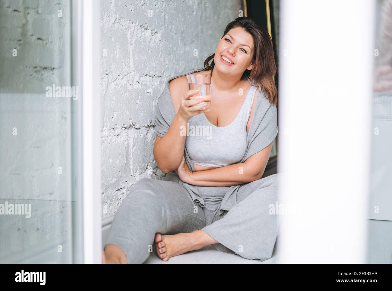 Beautiful happy emotional brunette young woman plus size body positive in comfortable sport wear with glass of water at home Stock Photo