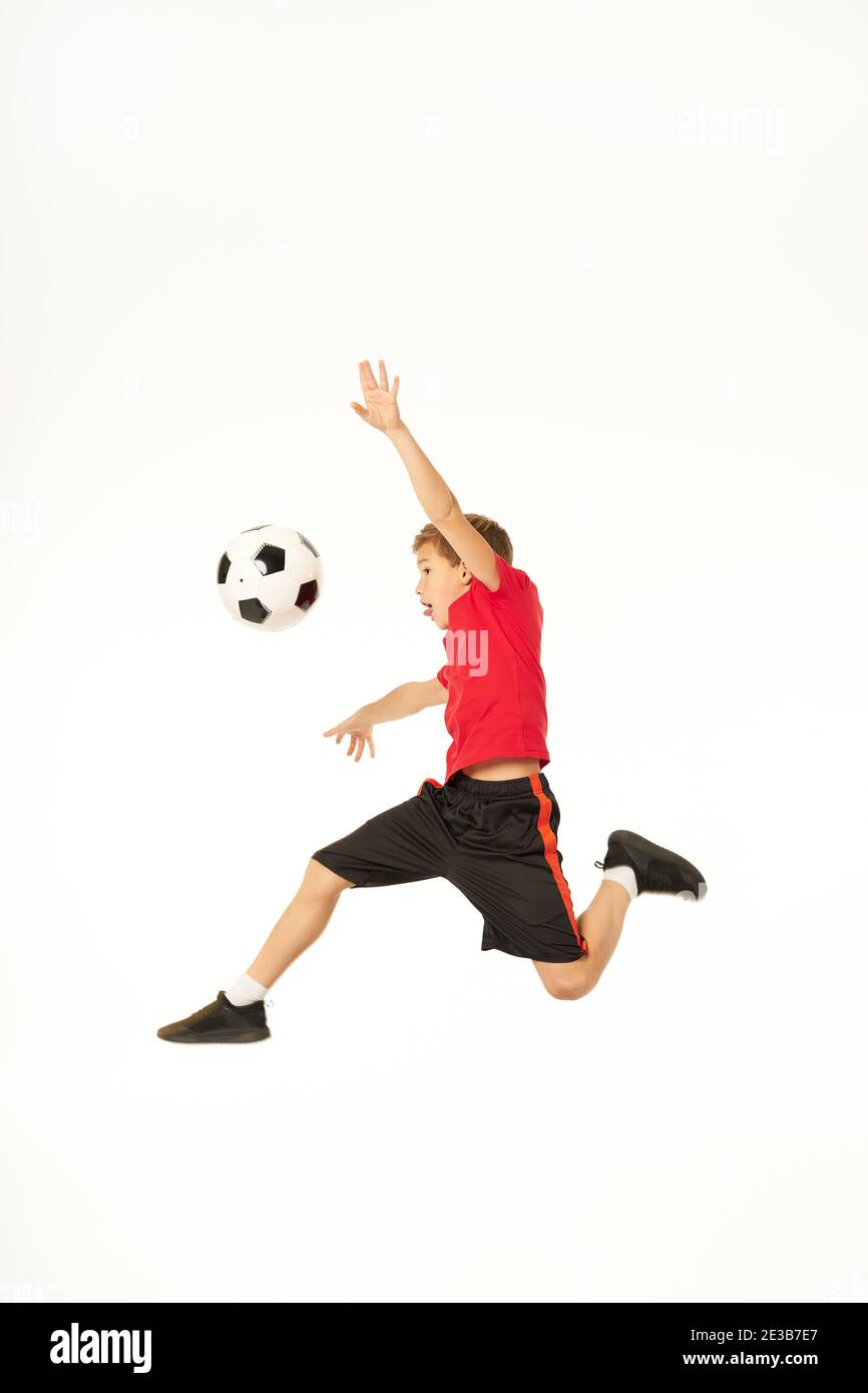 Cute boy football player looking at soccer ball and sticking out tongue while jumping in the air. Isolated on white background Stock Photo