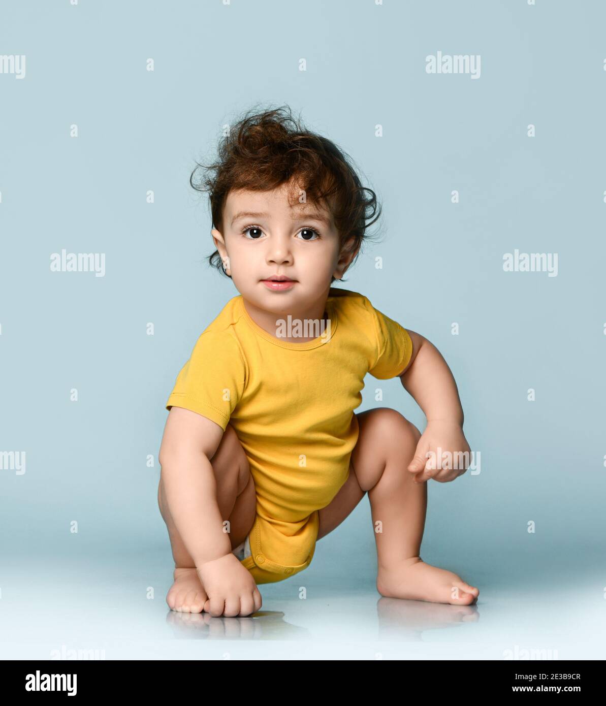 Cute smiling curly baby boy toddler in yellow comfortable jumpsuit sitting on floor and looking at camera Stock Photo