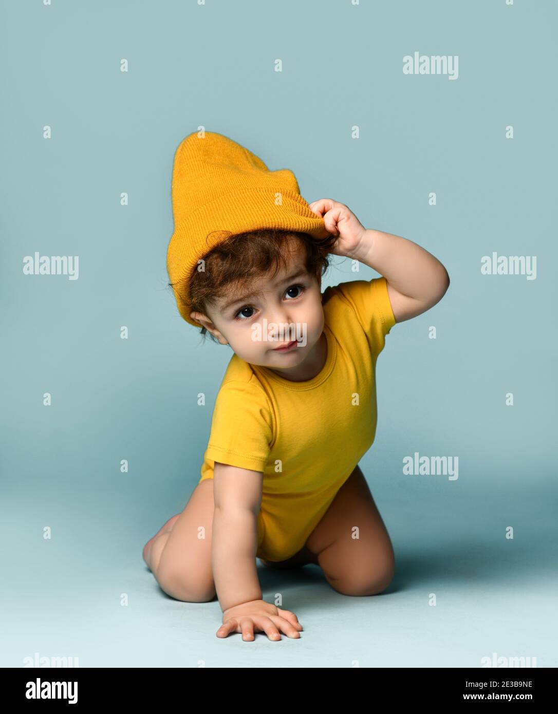 Small cute smiling curly baby boy toddler in yellow comfortable jumpsuit and hat crawling on floor Stock Photo