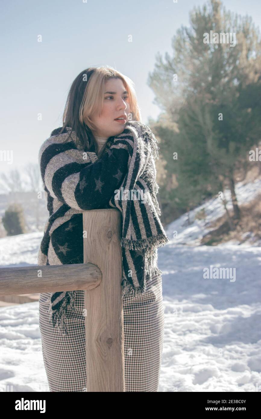 Vertical portrait of a beautiful woman with a huge scarf around her body from the cold leaning on a wooden pole being cold on a snowy winter day 2021. Stock Photo