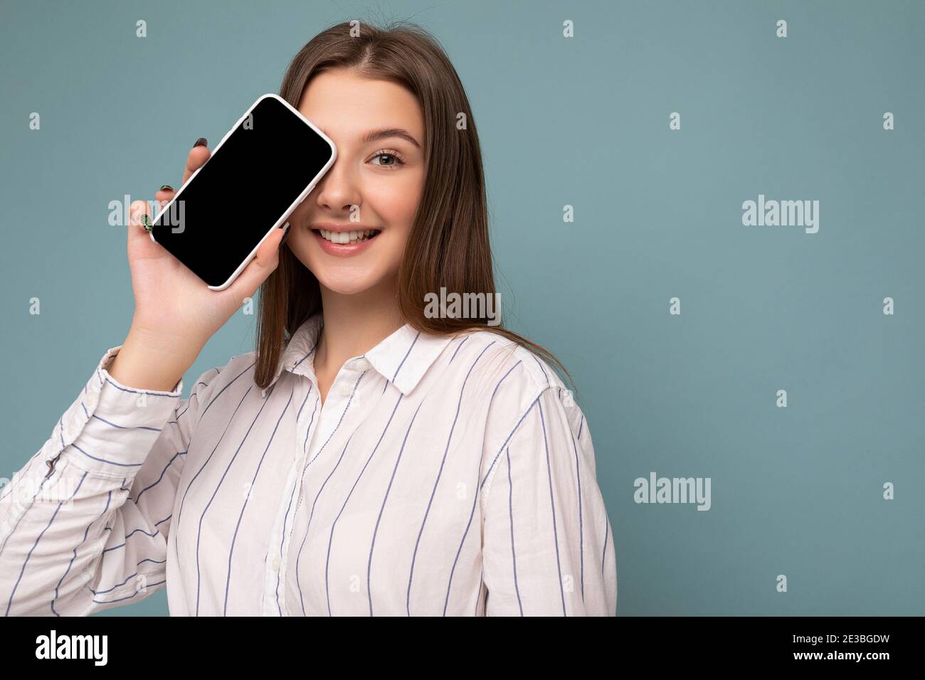 Closeup photo of Positive attractive young blonde woman wearing casual white shirt isolated over blue background wall holding smartphone and showing Stock Photo