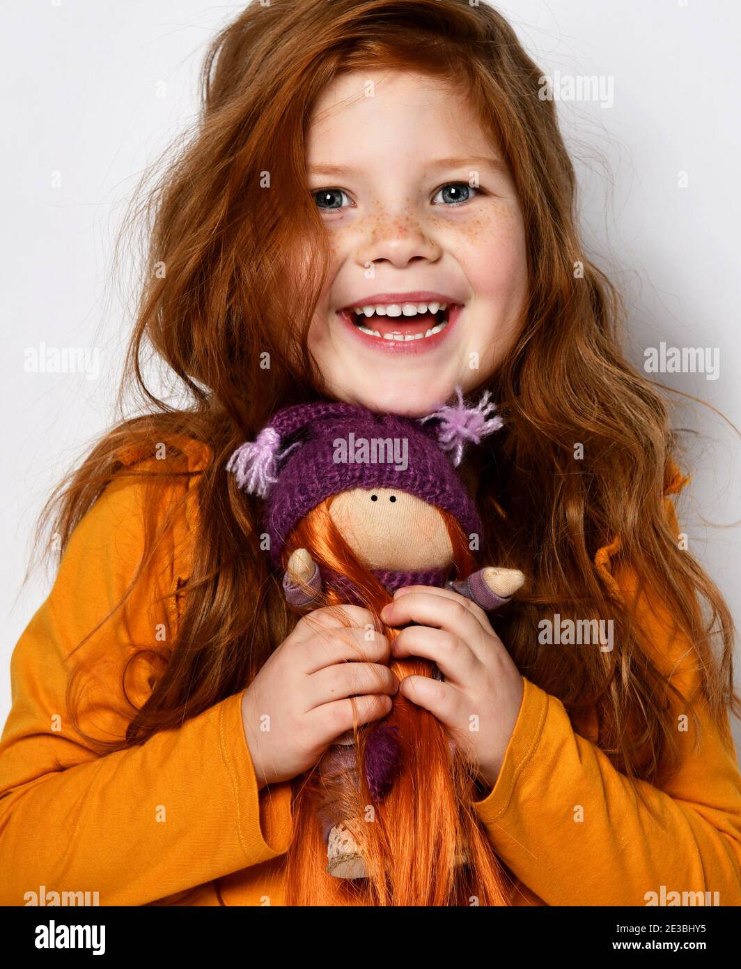 Little five-year-old red-haired girl smiles happily and holds a redhair doll in her hands in an orange sweater on white Stock Photo