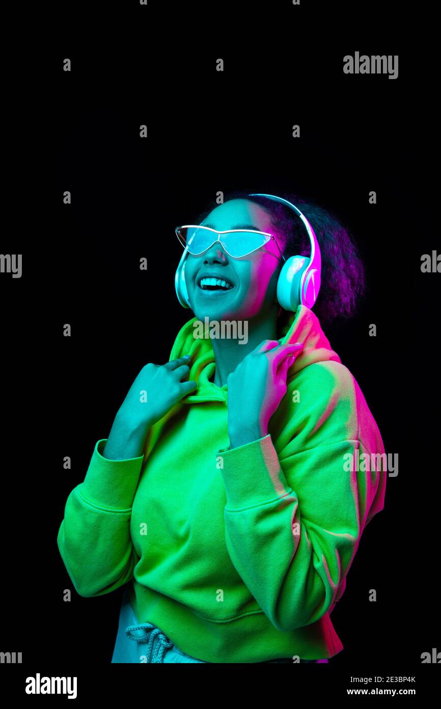 Dancer. African-american woman isolated on dark studio background in multicolored neon. Listening to music with headphones. Concept of human emotions, facial expression, sales, ad, fashion. Stock Photo