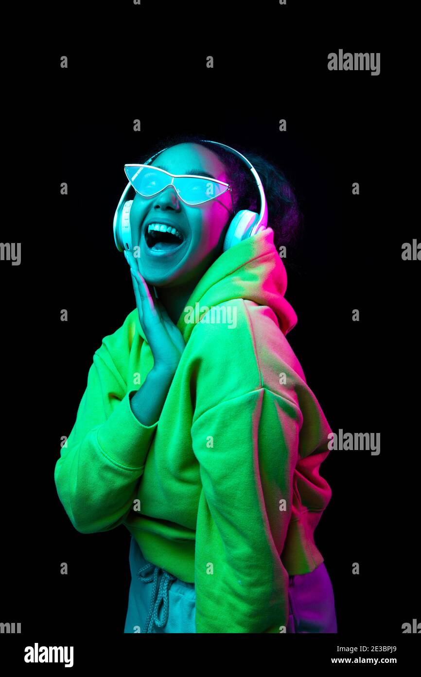 Astonished. African-american woman isolated on dark studio background in multicolored neon. Listening to music with headphones. Concept of human emotions, facial expression, sales, ad, fashion. Stock Photo