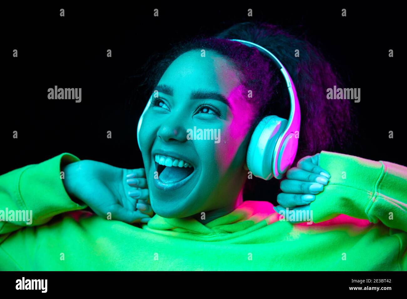 Laughting. African-american woman isolated on dark studio background in multicolored neon. Listening to music with headphones. Concept of human emotions, facial expression, sales, ad, fashion. Stock Photo
