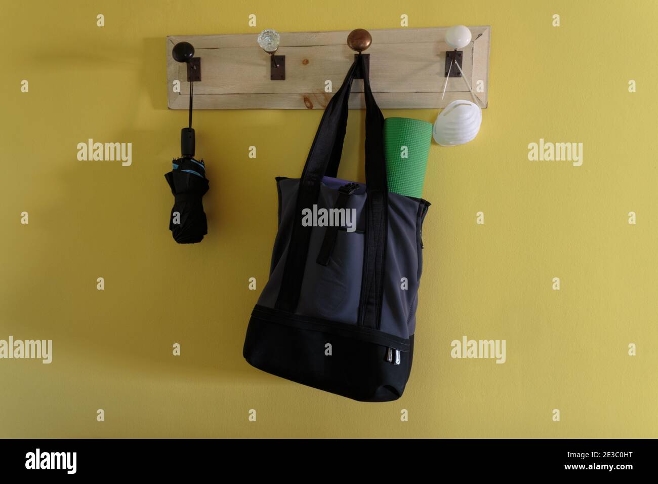 coat rack hanging on a bright yellow wall holding an umbrella, a sports bag with a rolled up yoga mat sticking out and a face mask Stock Photo