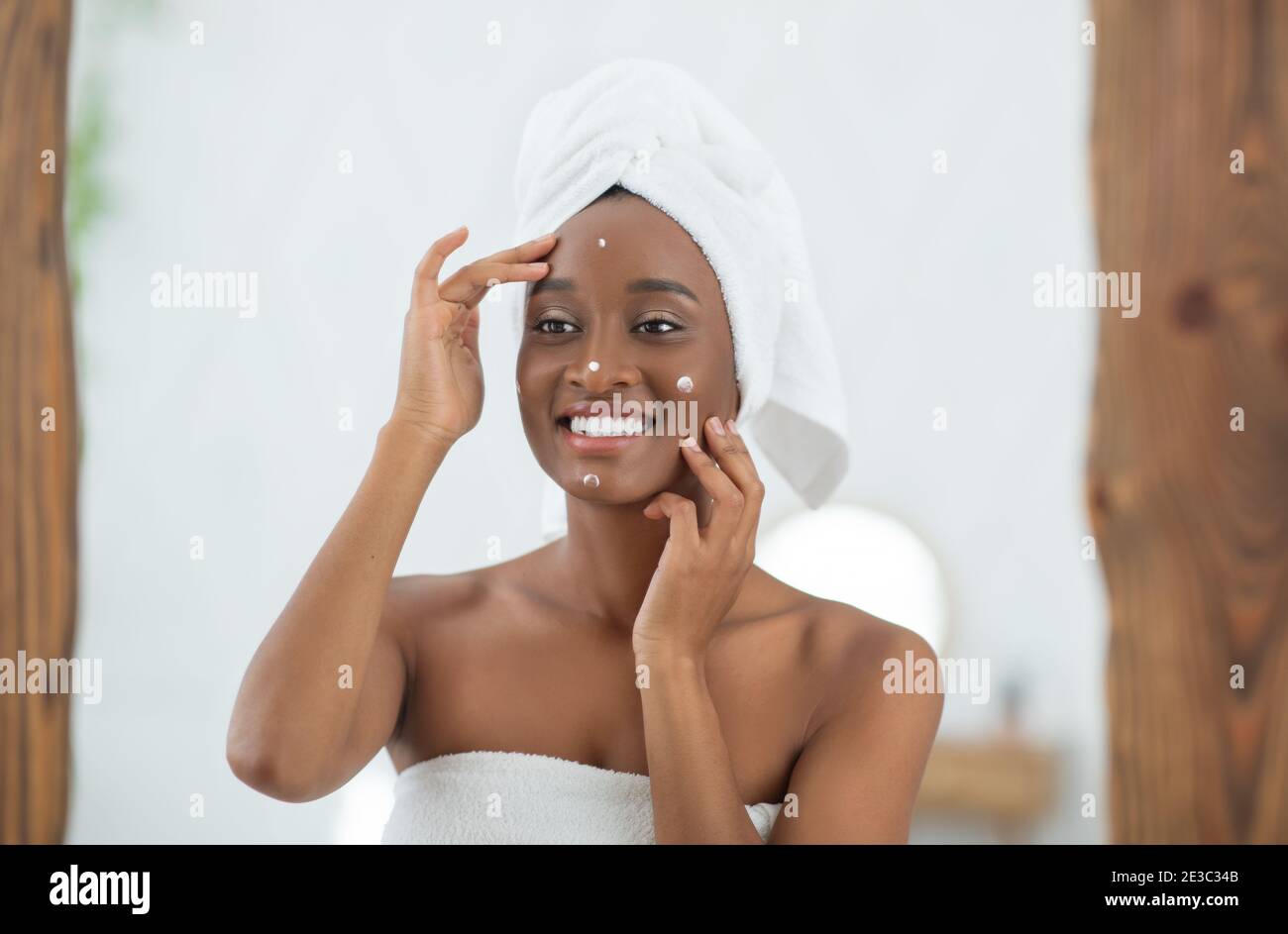 Happy african american lady with white towel on head looks in mirror and smears face with cream Stock Photo