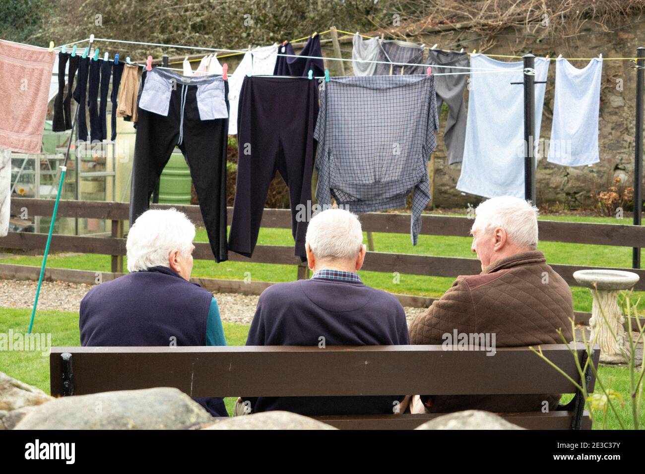 Three senior people with backs to camera sitting on bench with washing hanging in line in background Dunkeld Scotland Stock Photo
