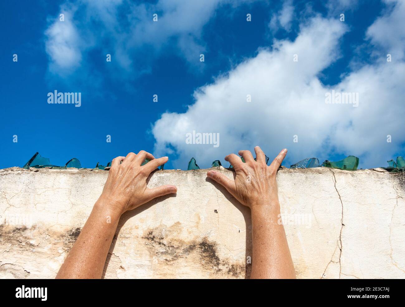 Hands on broken glass on top of wall with blue sky copy space. Escaping, trapped, prison, lockdown... concept. Stock Photo