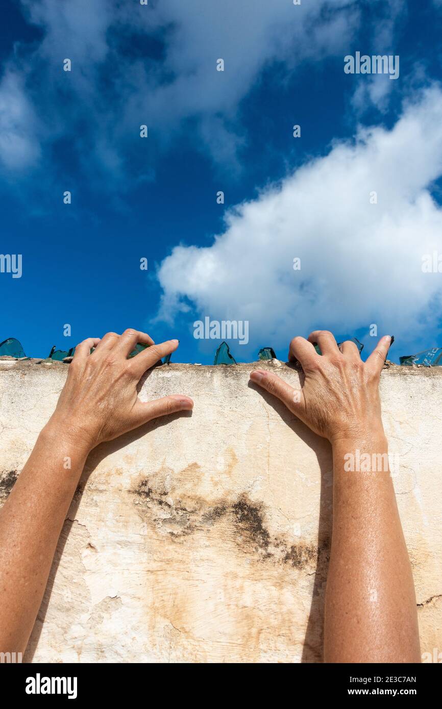 Hands on broken glass on top of wall with blue sky copy space. Escaping, trapped, prison, lockdown... concept. Stock Photo