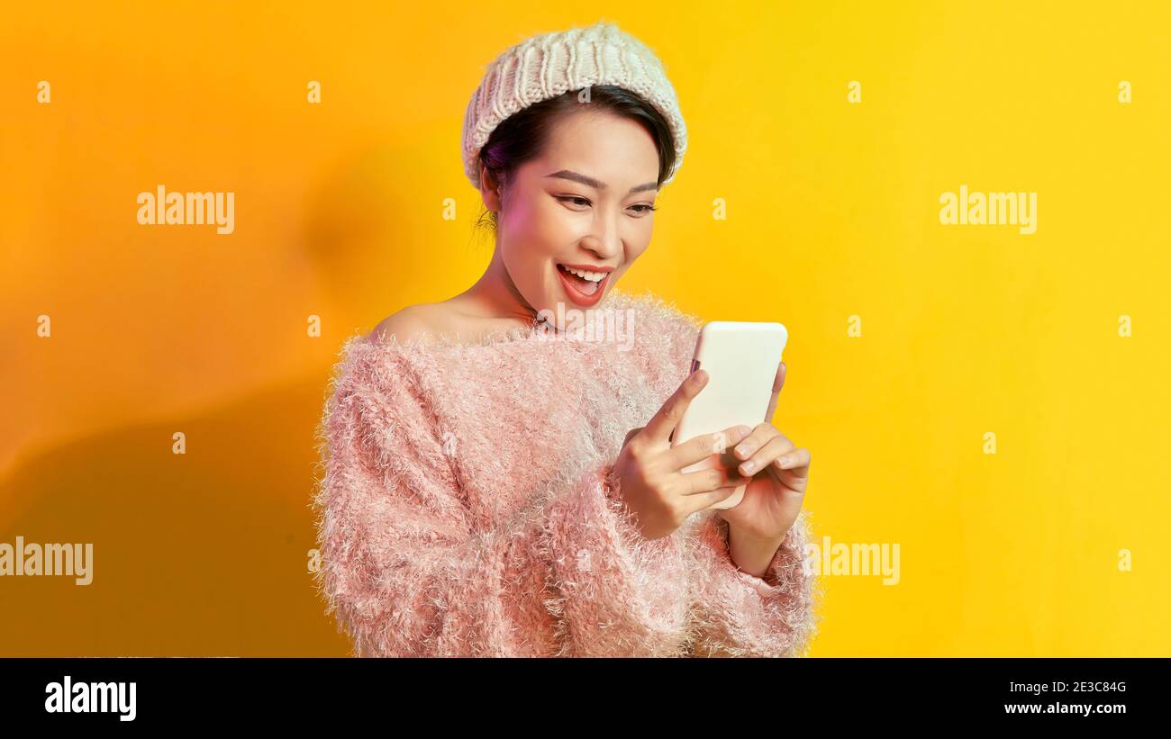 Pretty lady looks at telephone screen, chatting messages, wear warm casual hat, posing isolated over yellow background, Stock Photo