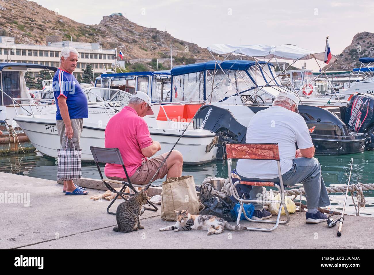 Sevastopol, Russia - January 7, 2020: Two male fishermen in casual clothes sit on folding chairs on the dock and fish. Several cute cats nearby are wa Stock Photo