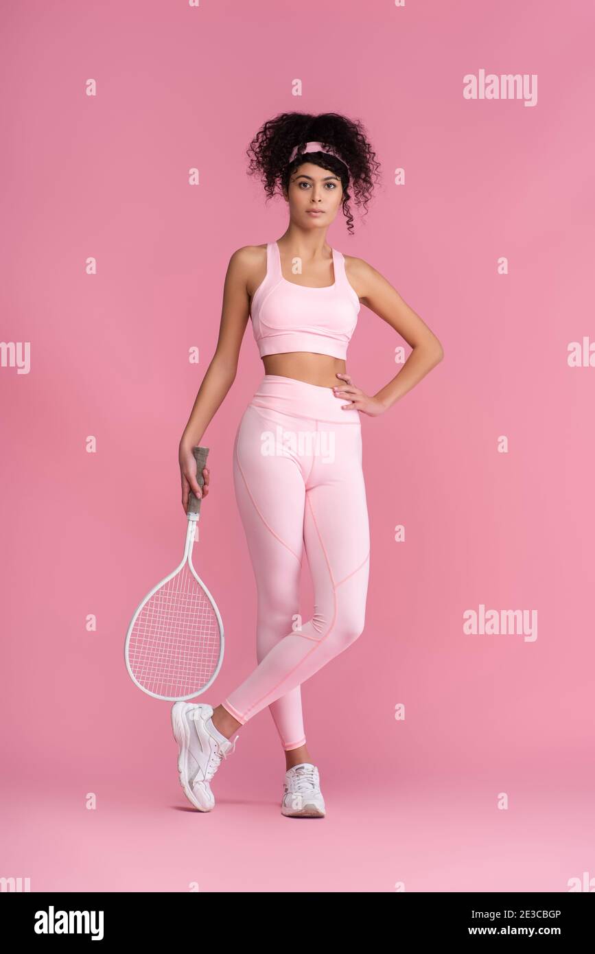 full length of curly woman in sportswear holding tennis racket while ...