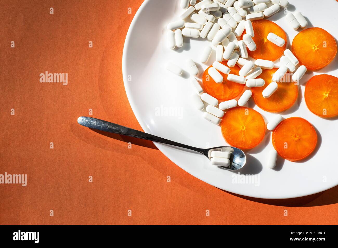 On an oval white plate - persimmon orange slices, capsules of mineral supplements and a spoon with a pill. Orange. Biohacking on health topics. Stock Photo