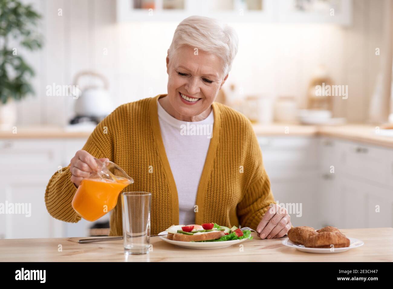 Attractive old lady pouring juice, having lunch in kitchen Stock Photo